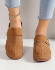 Tan mules on model feet with wide strap and metal buckle detail.