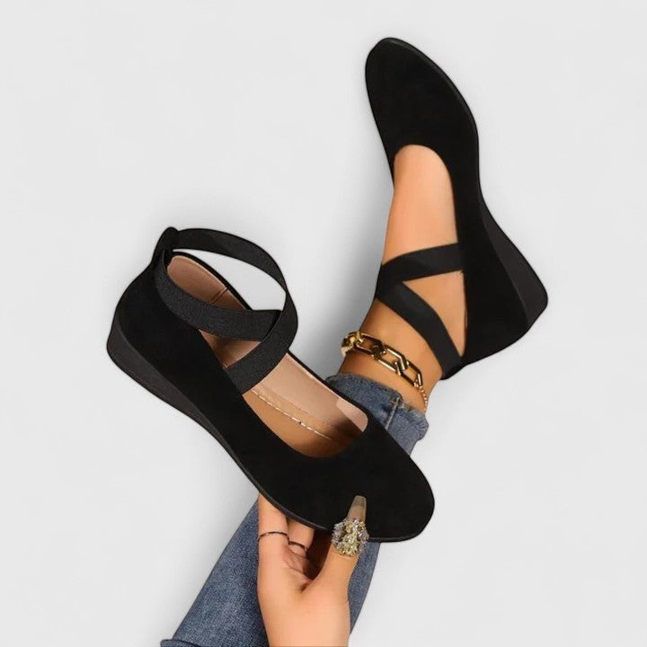 Black wedge ballet flats with ankle strap, crisscross upper, and gold bracelet detail.